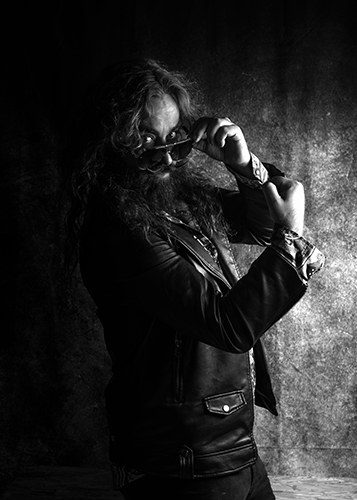 Black and white photograph of a man with long hair, beard and moustache wearing a leather jacket, flexing his arm and peering over his sunglasses